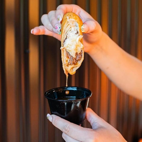 A hand holds a Tacos Number Juan birria taco, dripping consommé into a small black cup.
