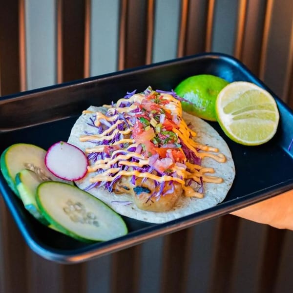 A Tacos Number Juan shrimp taco with purple cabbage, salsa, and sauce, served with lime and cucumber.