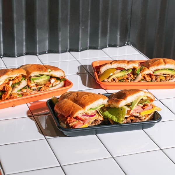 Three Tacos Number Juan tortas, filled with meat, avocado, and vegetables, on white tiled surface.